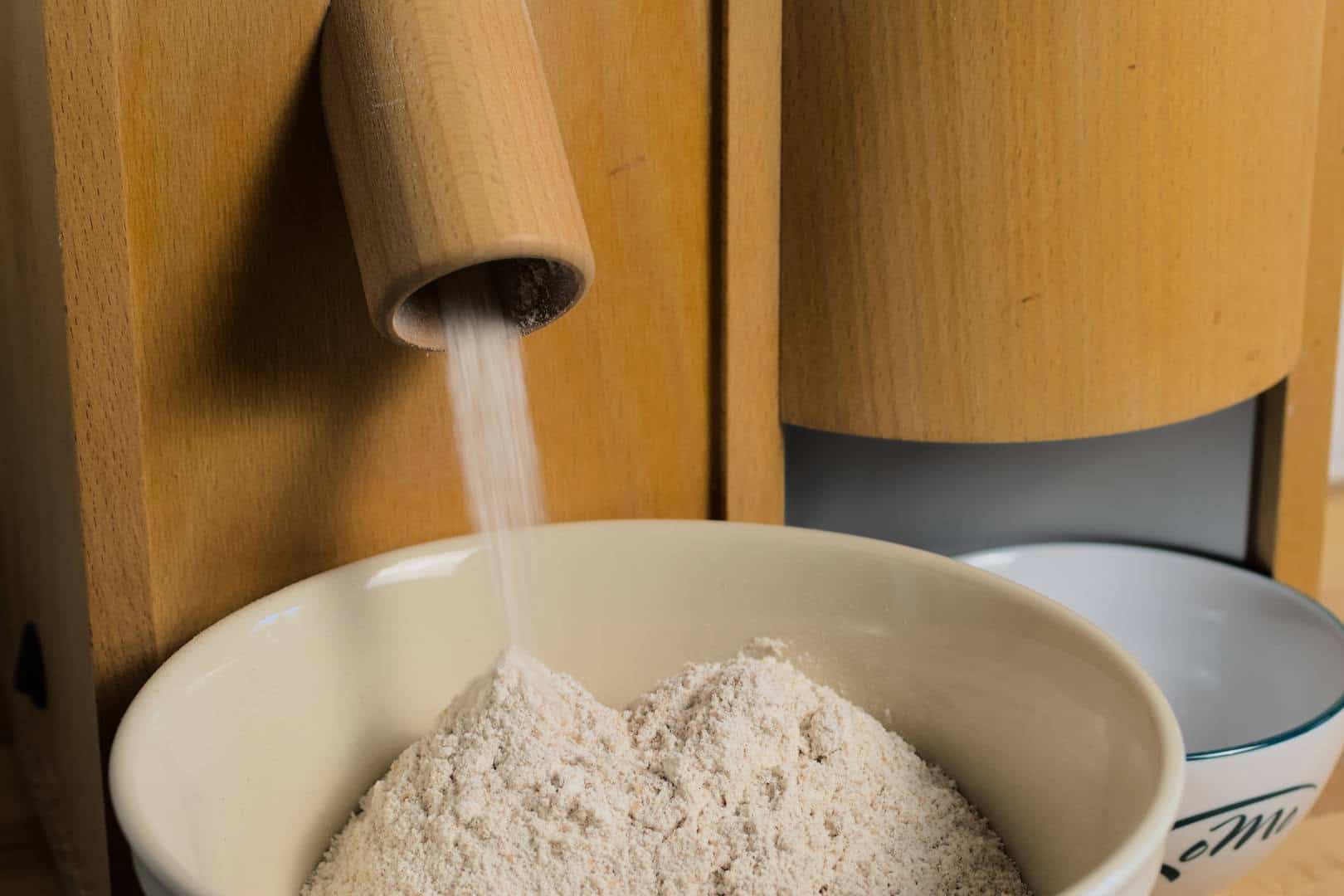 Closeup of freshly milled flour falling from the spout of a wooden grain mill into a white ceramic bowl