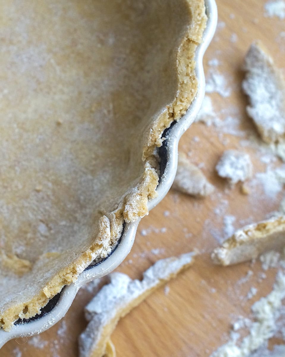 Closeup of sourdough pie crust in a tart pan, edges trimmed neatly with floury scraps of dough and a rolling pin next to it.