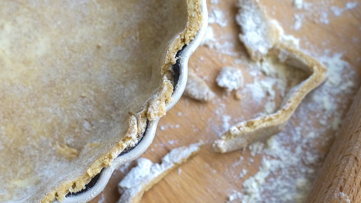 Closeup of sourdough pie crust in a tart pan, edges trimmed neatly with floury scraps of dough and a rolling pin next to it.