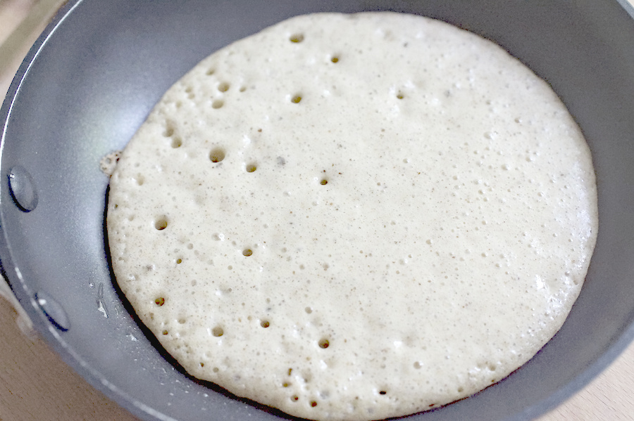 closeup ofp ancake batter in a frying pan. there are a lot of bubbles on top and the sides are just starting to set and brown.