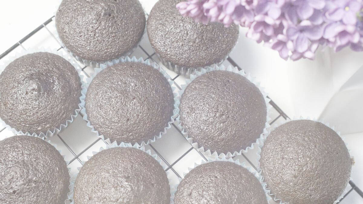 glossy black freshly milled vegan chocolate cupcakes in paper liners on black cooling rack, framed by a purple lilac