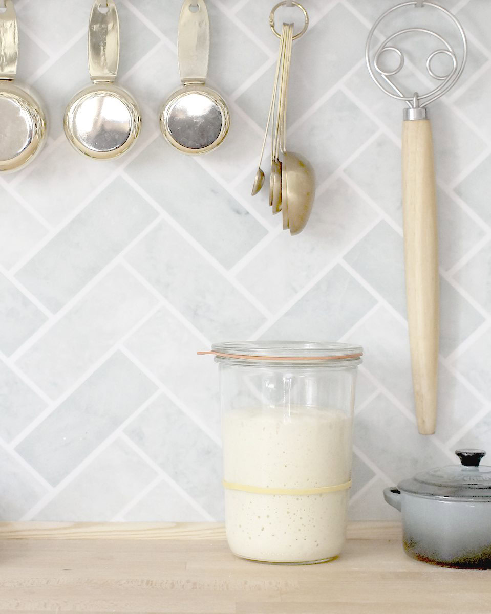 a warm wooden kitchen counter with a turquoise herringbone tile backsplash. Golden measuring cups and spoons hang from hooks above. On the counter sits a jar of sourdough starter and a small blue Le Creuset salt pot