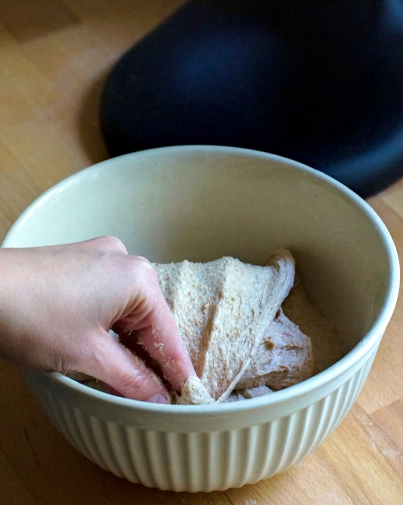 closeup of one hand folding a sourdough bread dough over on itself in a white ceramic bowl on a wooden counter