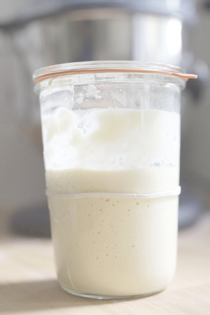 Closeup of a large jar full of deflated sourdough discard on a wooden kitchen counter with a black stand mixer in the background.
