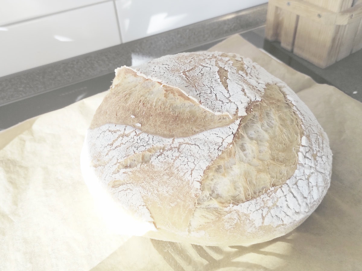 Crusty round loaf of sourdough bread made without all refined soft wheat flour, resting on parchment paper.