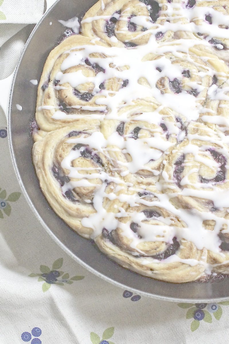 Closeup of Blueberry Lemon Sourdough Sweet Rolls, showing jammy berry swirls wrapped in a golden sourdough discard dough, topped with ribbons of white lemon glaze