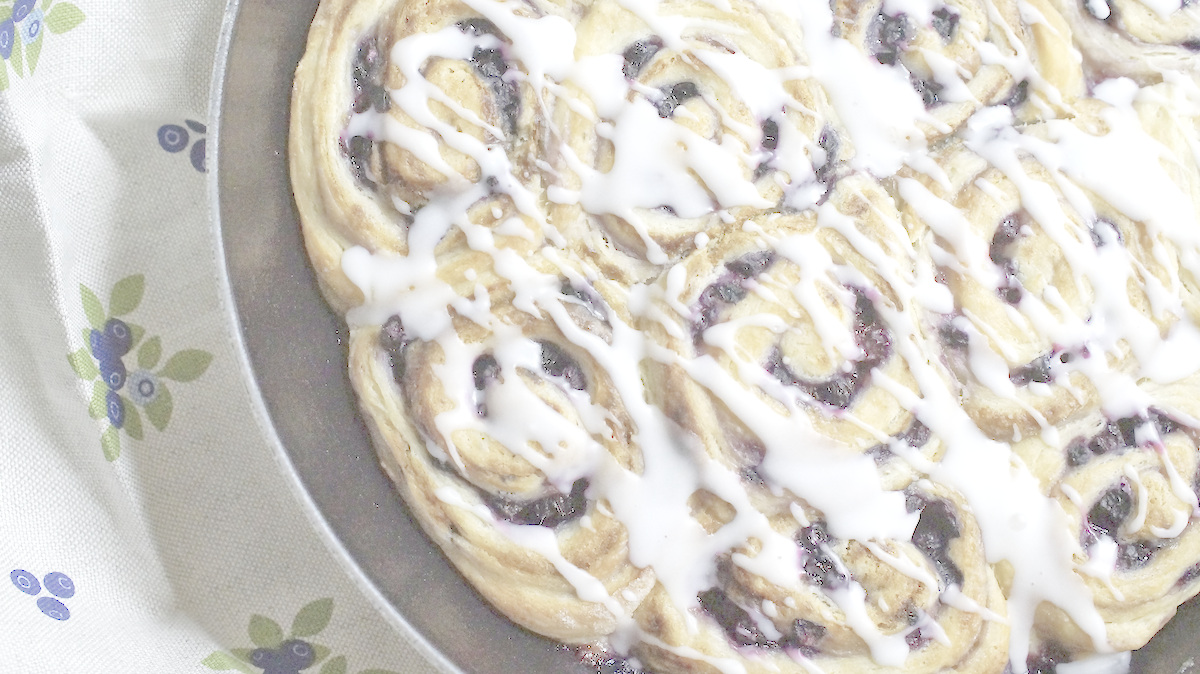 Closeup of Blueberry Lemon Sourdough Sweet Rolls, showing jammy berry swirls wrapped in a golden sourdough discard dough, topped with ribbons of white lemon glaze