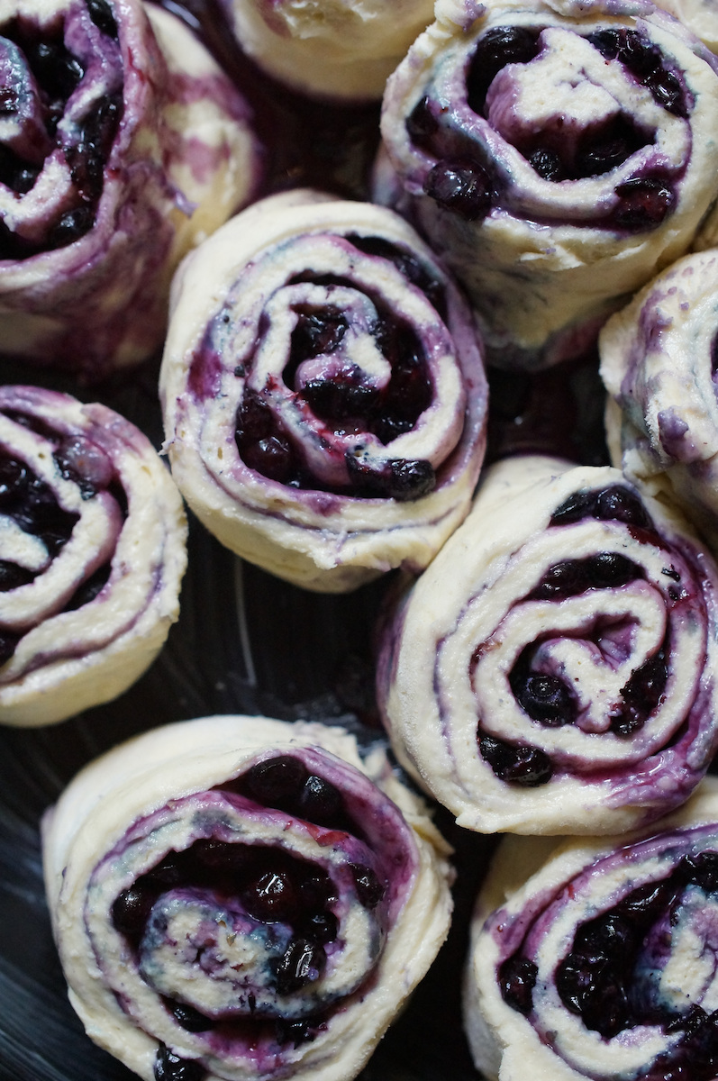 Unbaked blueberry sourdough sweet rolls, rolled up tightly and sitting side by side in a black cast iron pan.