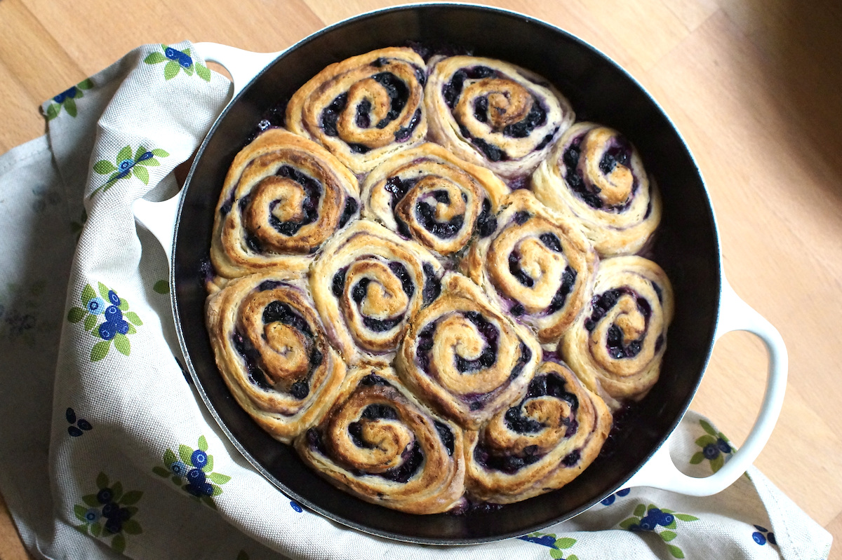 Golden baked unglazed Sourdough Berry Sweet Rolls, in a black cast iron pan with white handles, sitting on a warm wooden kitchen counter, surrounded by a linen towel with a playful blueberry pattern. The dark swirls of blueberry filling contrast perfectly with the golden dough.