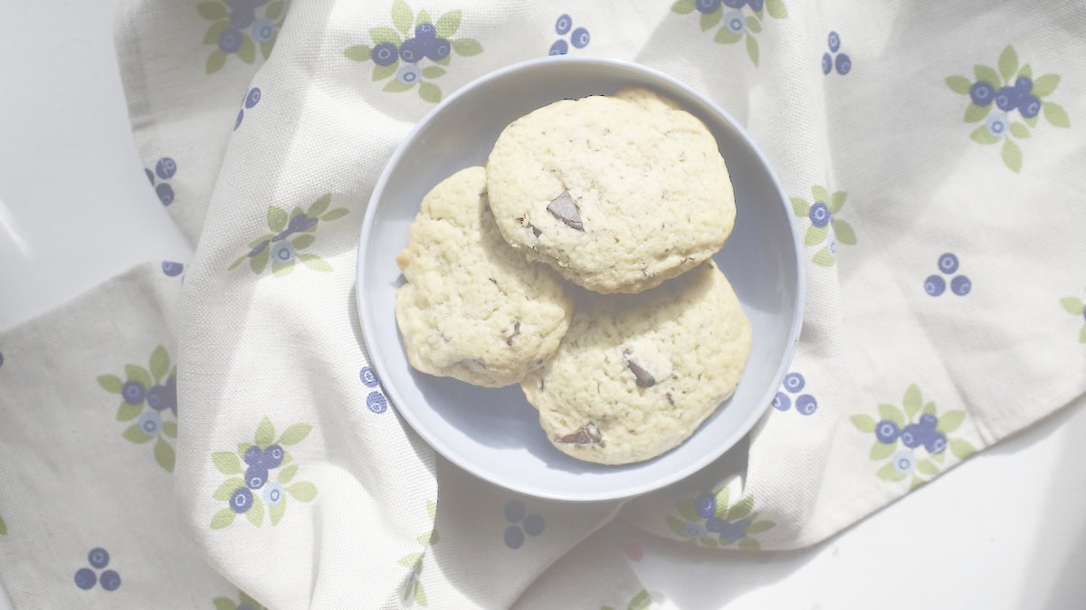 A blue bowl of golden Sourdough discard chocolate chip cookies sitting on a blueberry-patterned linen tea towel in golden sunlight.