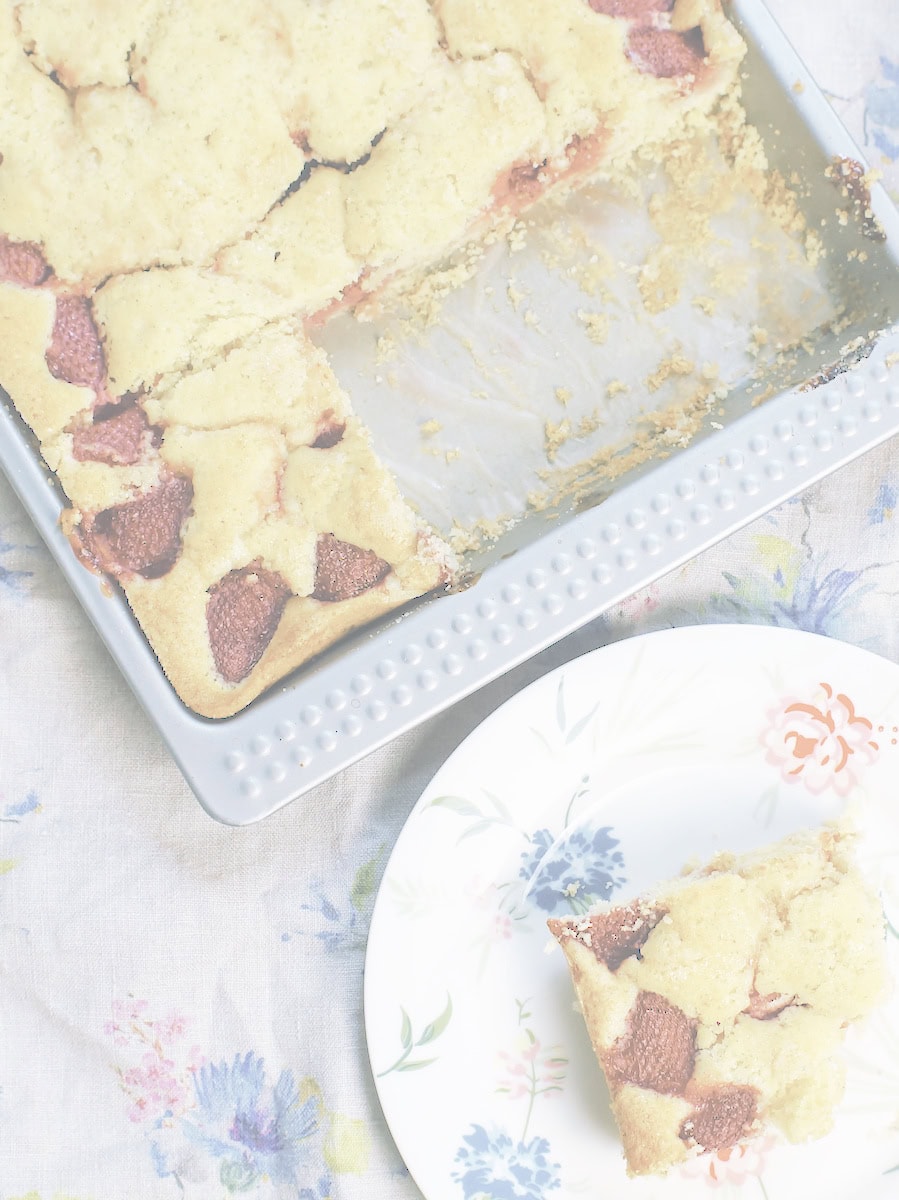 A slice of golden strawberry sourdough sheet cake on a plate, next to the pan, all set on a vintage floral tablecloth.