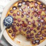 Overhead shot of golden baked sourdough discard clafoutis, in a ceramic pie dish, being spooned out with a blue, patterned ceramic spoon. Dark pockets of baked cherries are dusted with powdered sugar.