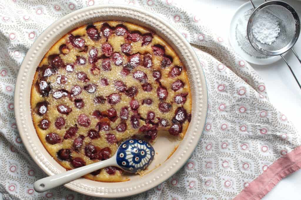 Overhead shot of golden baked sourdough discard clafoutis, in a ceramic pie dish, being spooned out with a blue, patterned ceramic spoon. Dark pockets of baked cherries are dusted with powdered sugar.