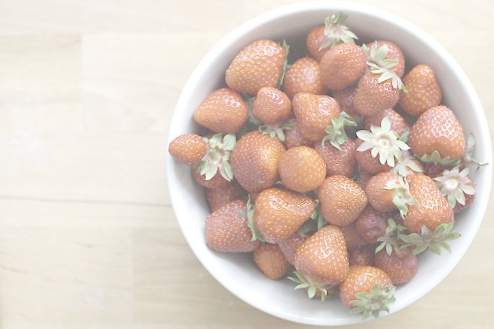 Bright red strawberries with green leaves in a white bowl on a wooden kitchen counter.