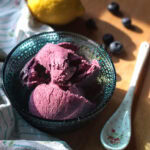 Purple blueberry ricotta ice cream served in a teal glass bowl, surrounded by fresh blueberries, a floral spoon, and a draped tea towel, all glowing in soft sunlight.