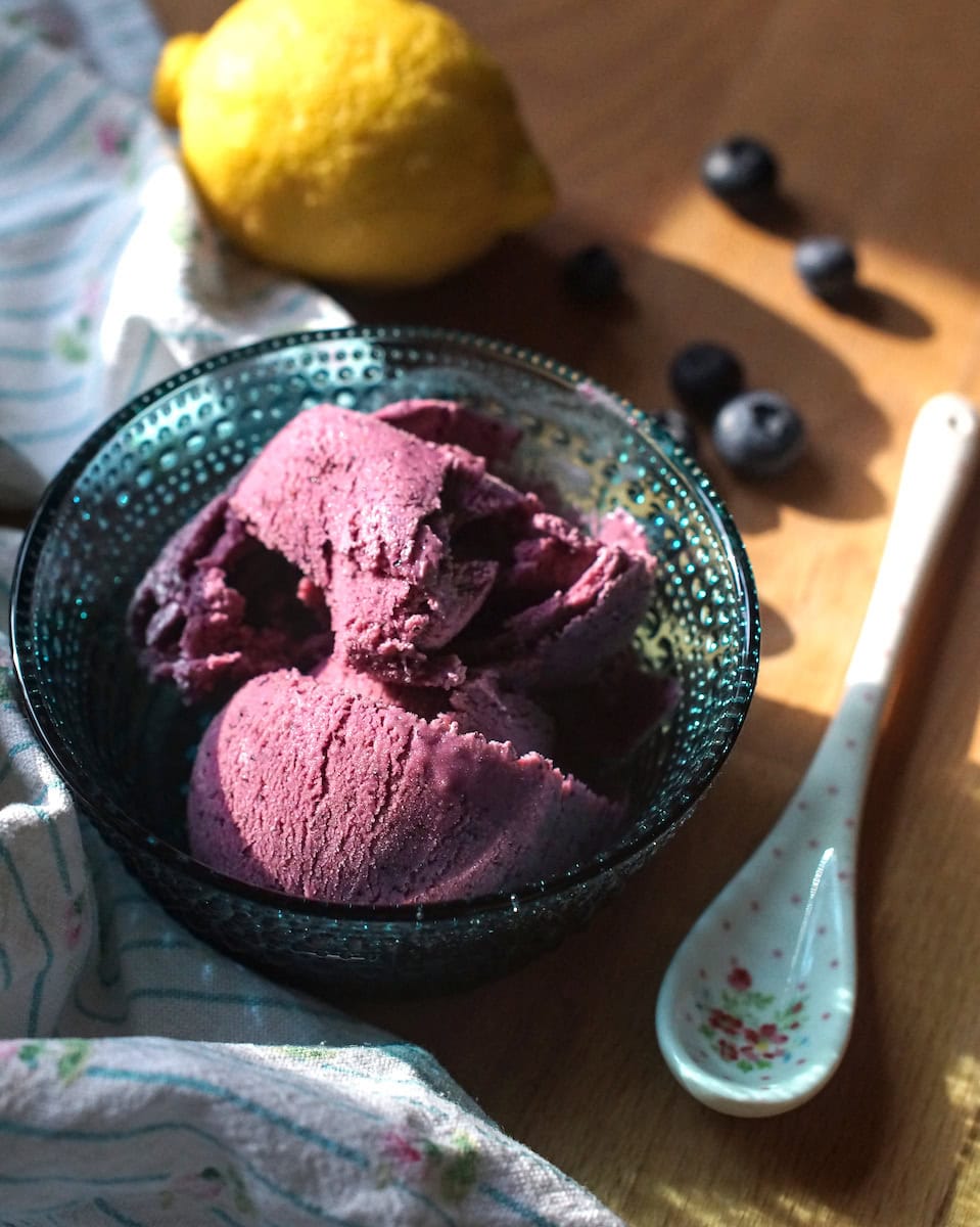 Purple blueberry ricotta ice cream served in a teal glass bowl, surrounded by fresh blueberries, a floral spoon, and a draped tea towel, all glowing in soft sunlight.