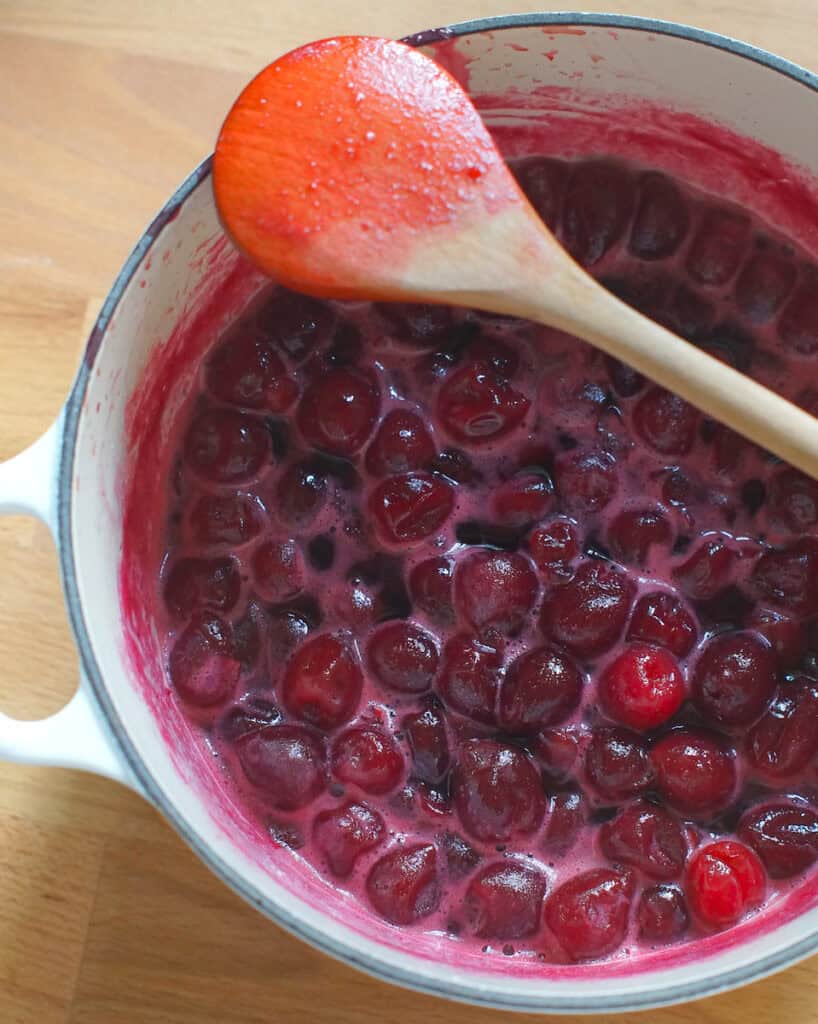 Cherry cardamom compote bubbling and foaming gently in the pot, with a wooden spoon resting across the rim