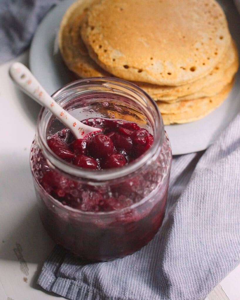 Open jar of glossy cherry cardamom compote with a floral ceramic spoon inside, set on a table with a plate of stacked pancakes blurred in the background and a blue-and-white striped tea towel nearby