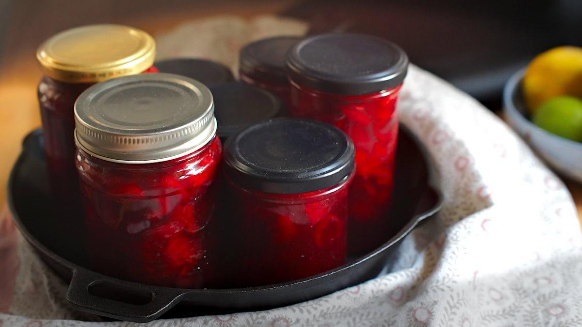 Jars full of easy homemade cherry lime jam shining in golden sunlight. Jars sit in a black cast iron pan wrapped in a patterned tea towel.