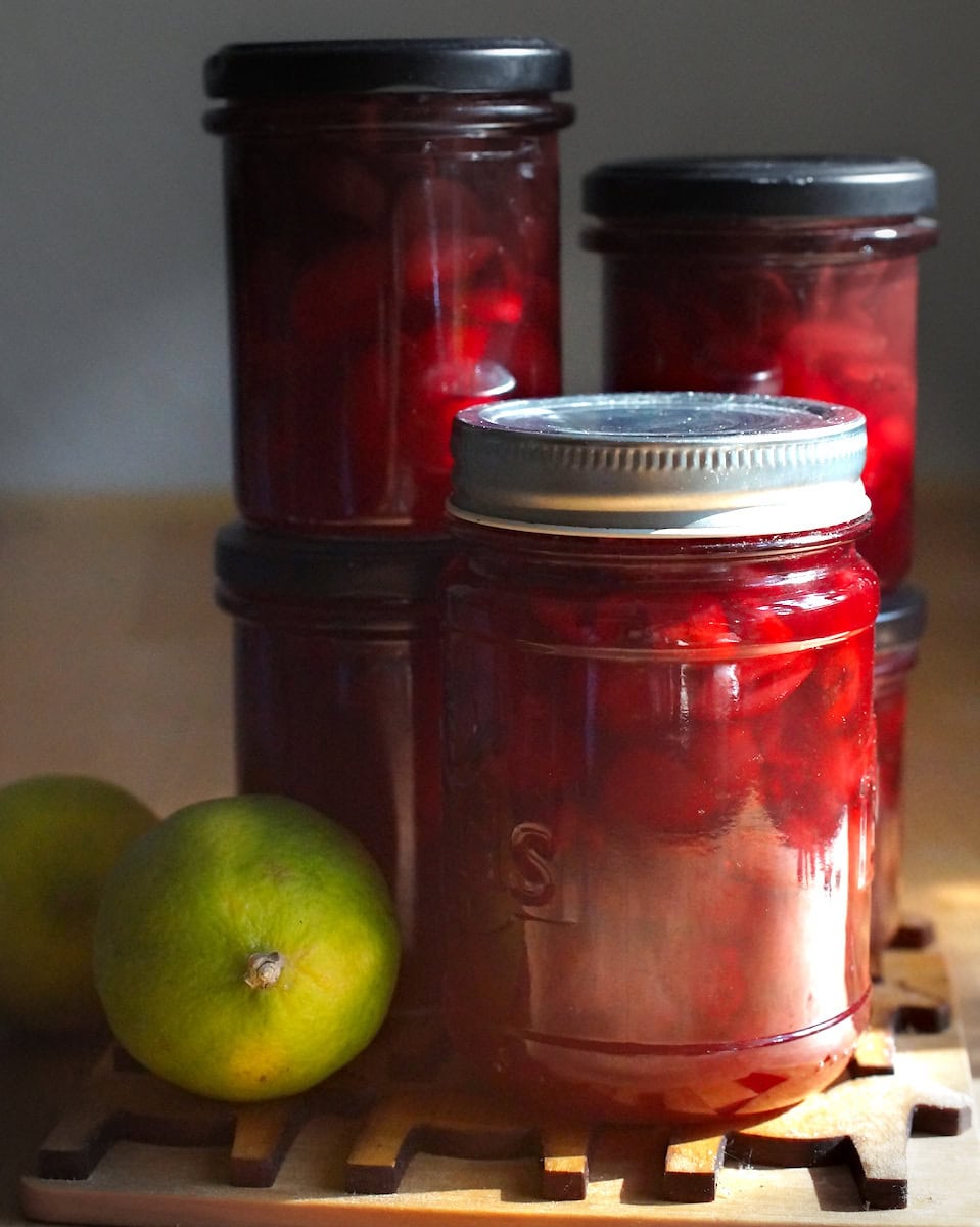 Jars full of easy homemade cherry lime jam shining in golden sunlight. Jars sit on a wooden caster along with the green limes.