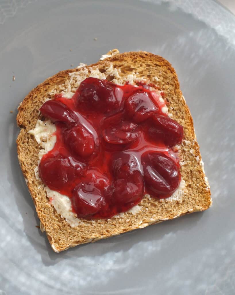 Easy homemade cherry lime jam spread on a golden toasted slice of sandwich bread, sitting on a pale blue plate in streaky sunlight.