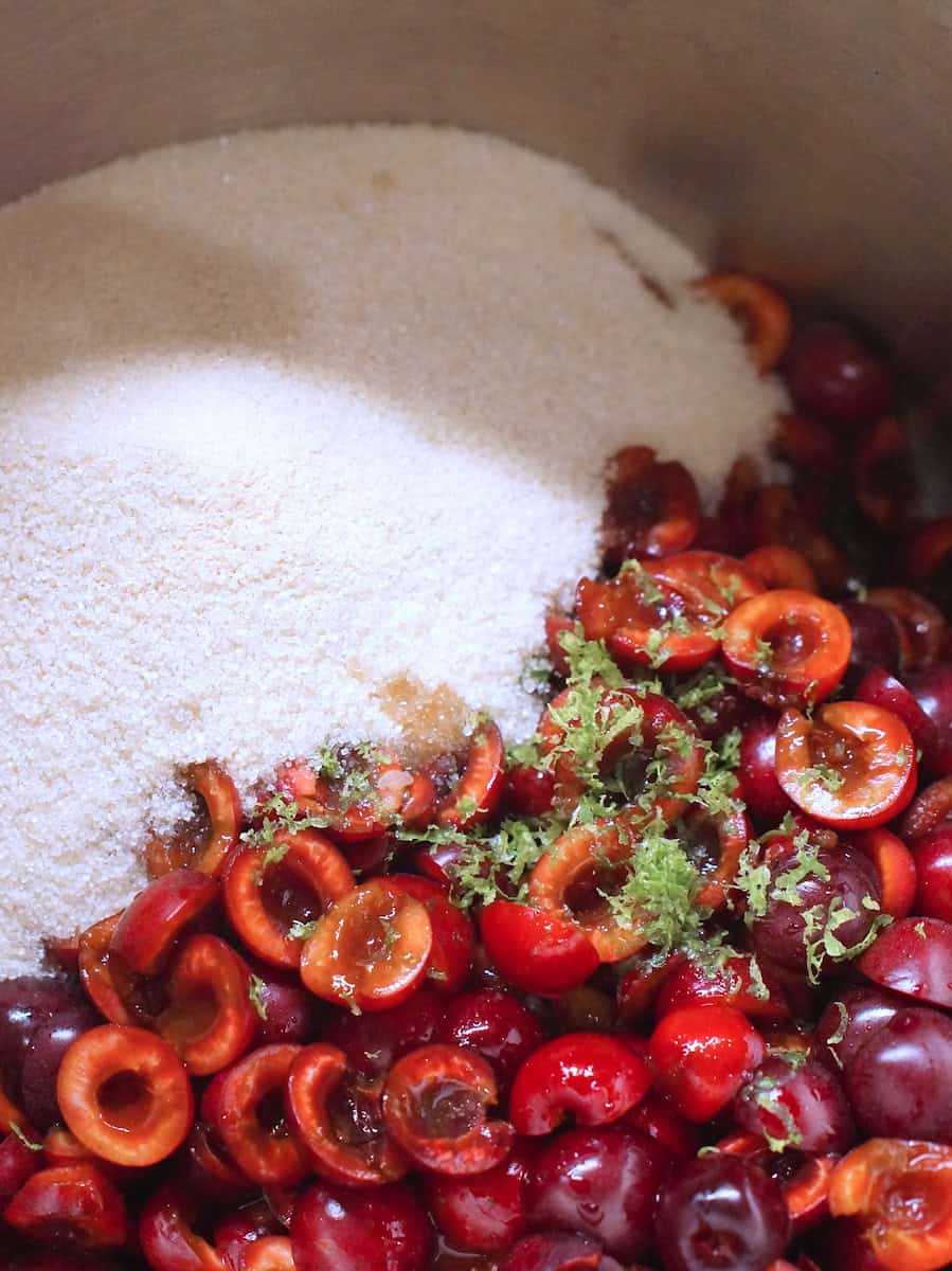 Bright white sugar, dark red cherries and vibrant green lime zest in a large silver pot, ready to be cooked into jam.