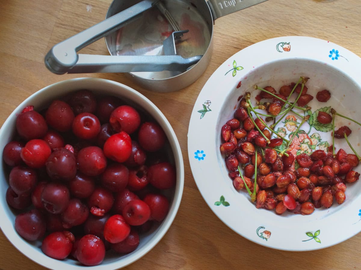 A bowl of vibrant, dark red cherries sits next to a bowl with the collected pits and stems. A measuring cup and cherry pitter sit on the side.