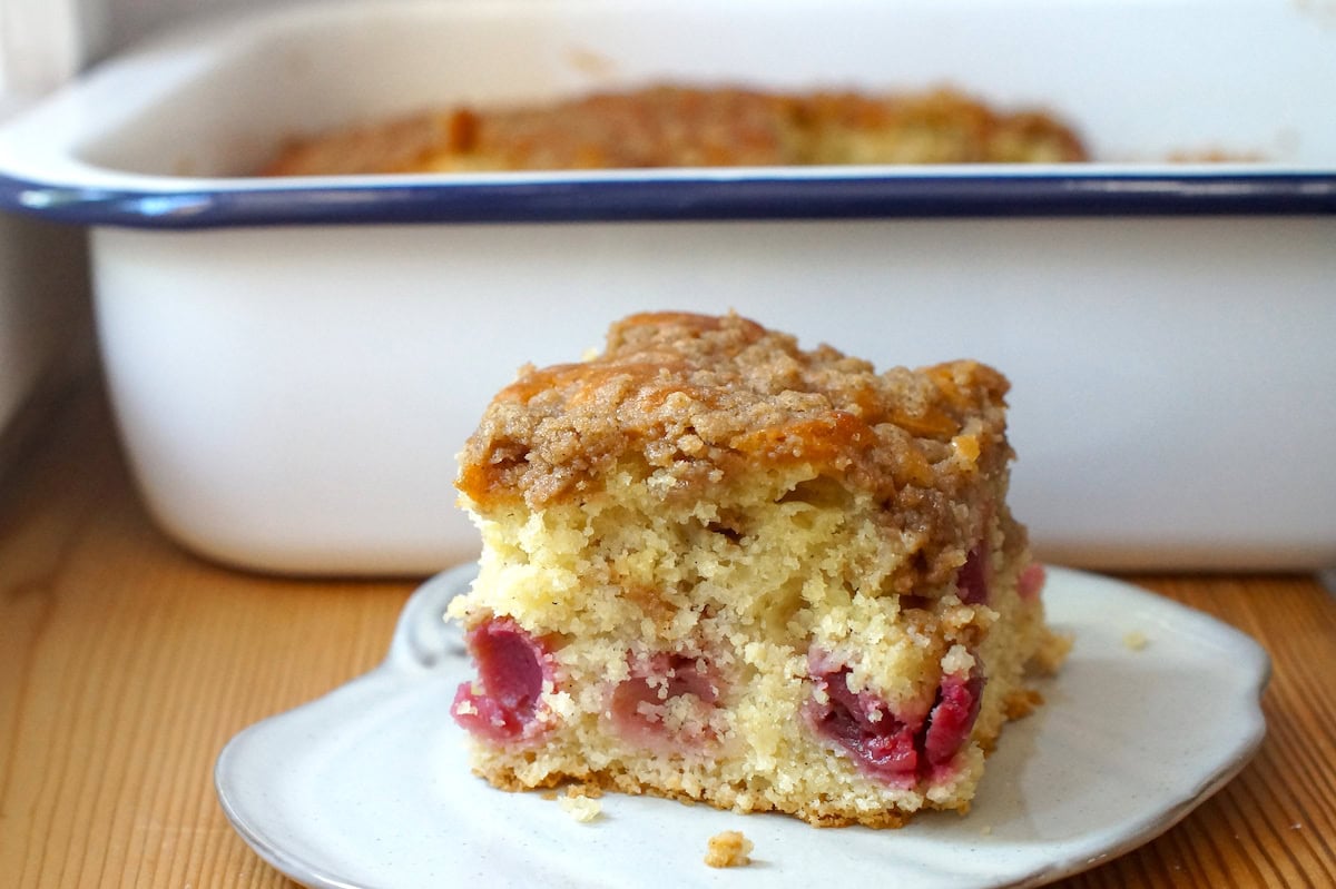 A square of sourdough discard cherry coffee cake on a small white plate, in front of a white baking pan with a dark blue rim.