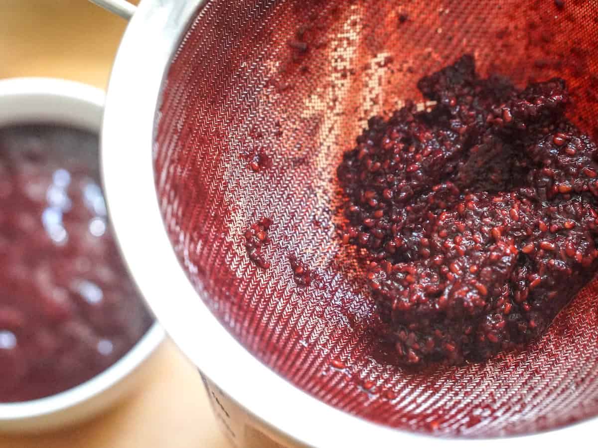 Deep red blackberry seeds in a fine mesh strainer, with a bowl of silky blackberry puree in the background.