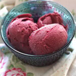 Three scoops of creamy blackberry Greek yogurt ice cream, sitting in a blue glass bowl, set out on a floral tea towel.