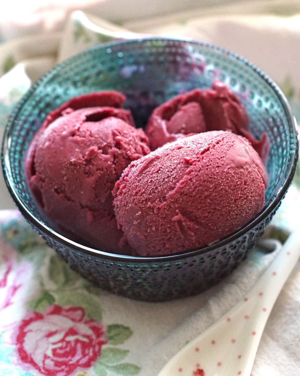 Three scoops of creamy blackberry Greek yogurt ice cream, sitting in a blue glass bowl, set out on a floral tea towel.
