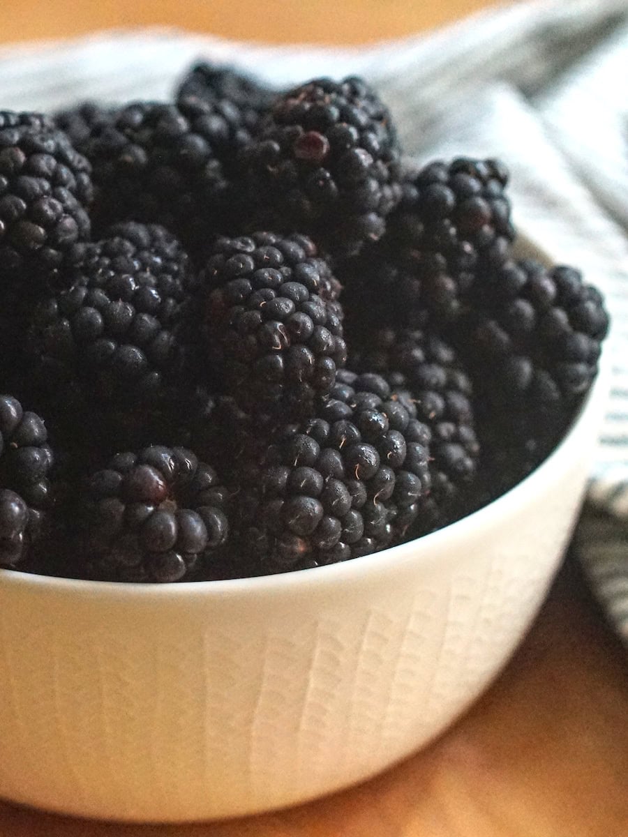 Closeup of glossy blackberries in a patterned white bowl, draped with a striped tea towel.