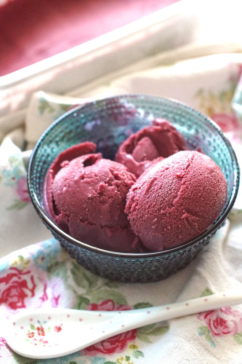 Three scoops of creamy blackberry Greek yogurt ice cream, sitting in a blue glass bowl, set out on a floral tea towel.