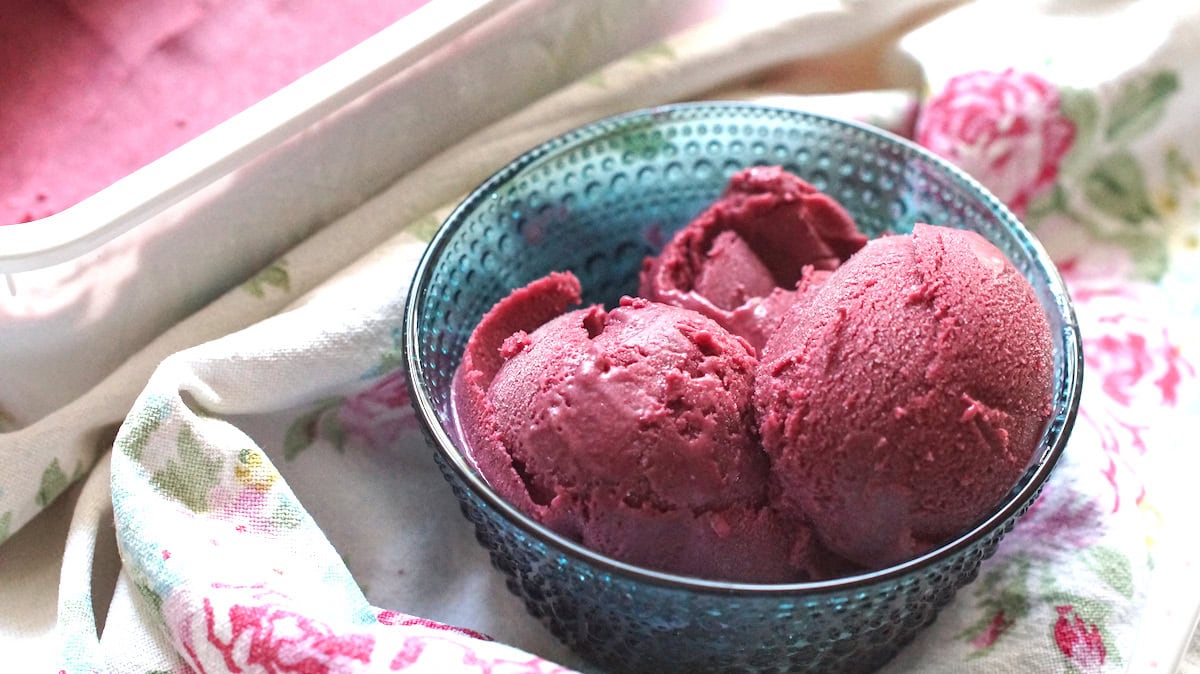 Three scoops of creamy blackberry Greek yogurt ice cream, sitting in a blue glass bowl, set out on a floral tea towel.