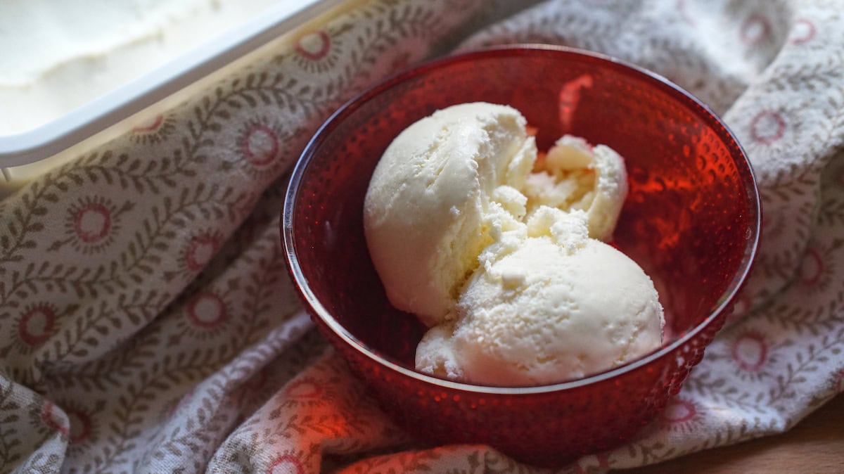 Two scoops of honey buttermilk ice cream are served in a red glass bowl, wrapped in a tea towel with red details. In the background you can see the freezer container full of buttermilk ice cream glistening in the sunlight.