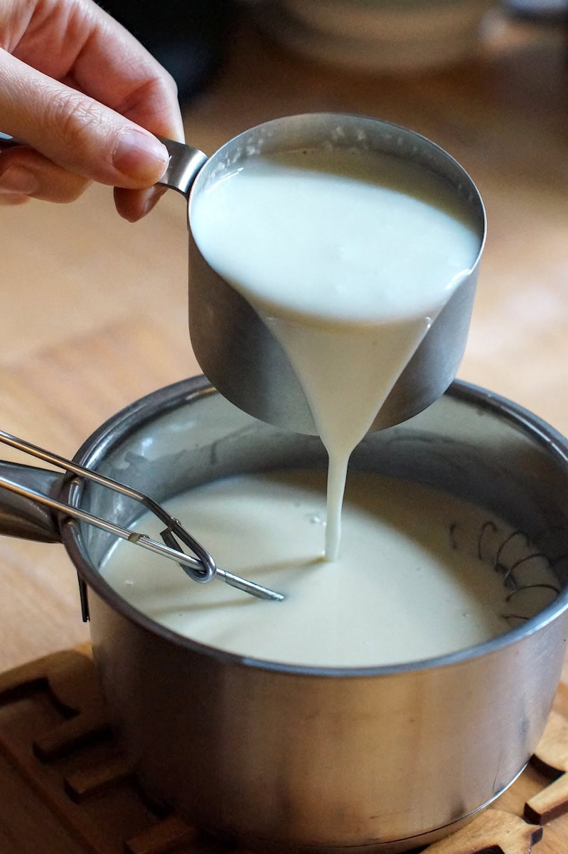 A cup of white buttermilk is being poured into a small saucepan with honey sweetened cream.