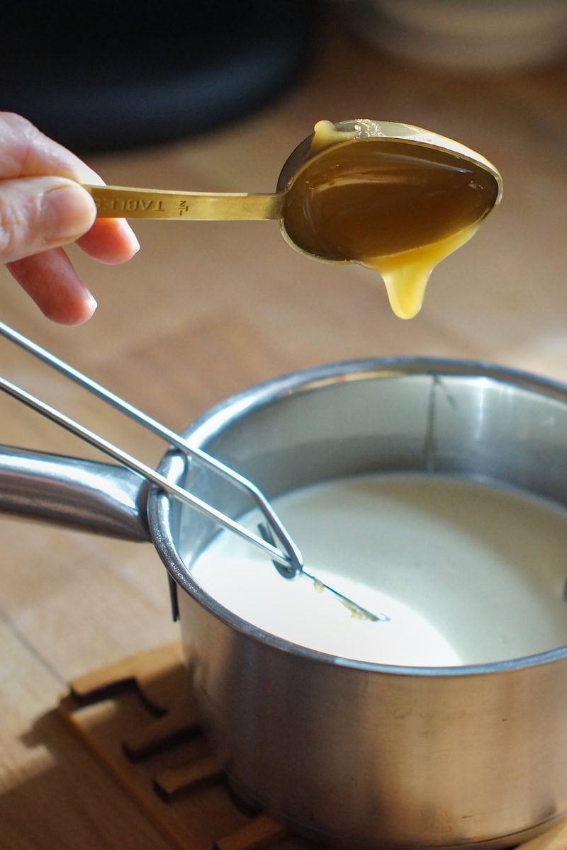 A spoonful of golden honey is dripping into a small saucepan filled with sweet cream.
