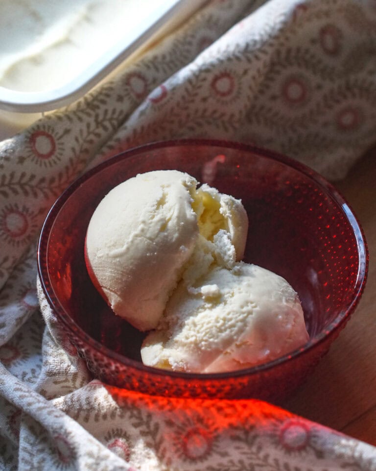 Two scoops of honey buttermilk ice cream are served in a red glass bowl, wrapped in a tea towel with red details. In the background you can see the freezer container full of buttermilk ice cream glistening in the sunlight.
