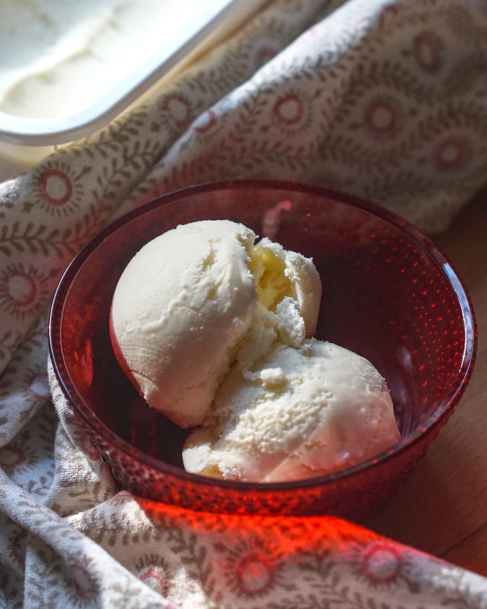 Two scoops of honey buttermilk ice cream are served in a red glass bowl, wrapped in a tea towel with red details. In the background you can see the freezer container full of buttermilk ice cream glistening in the sunlight.