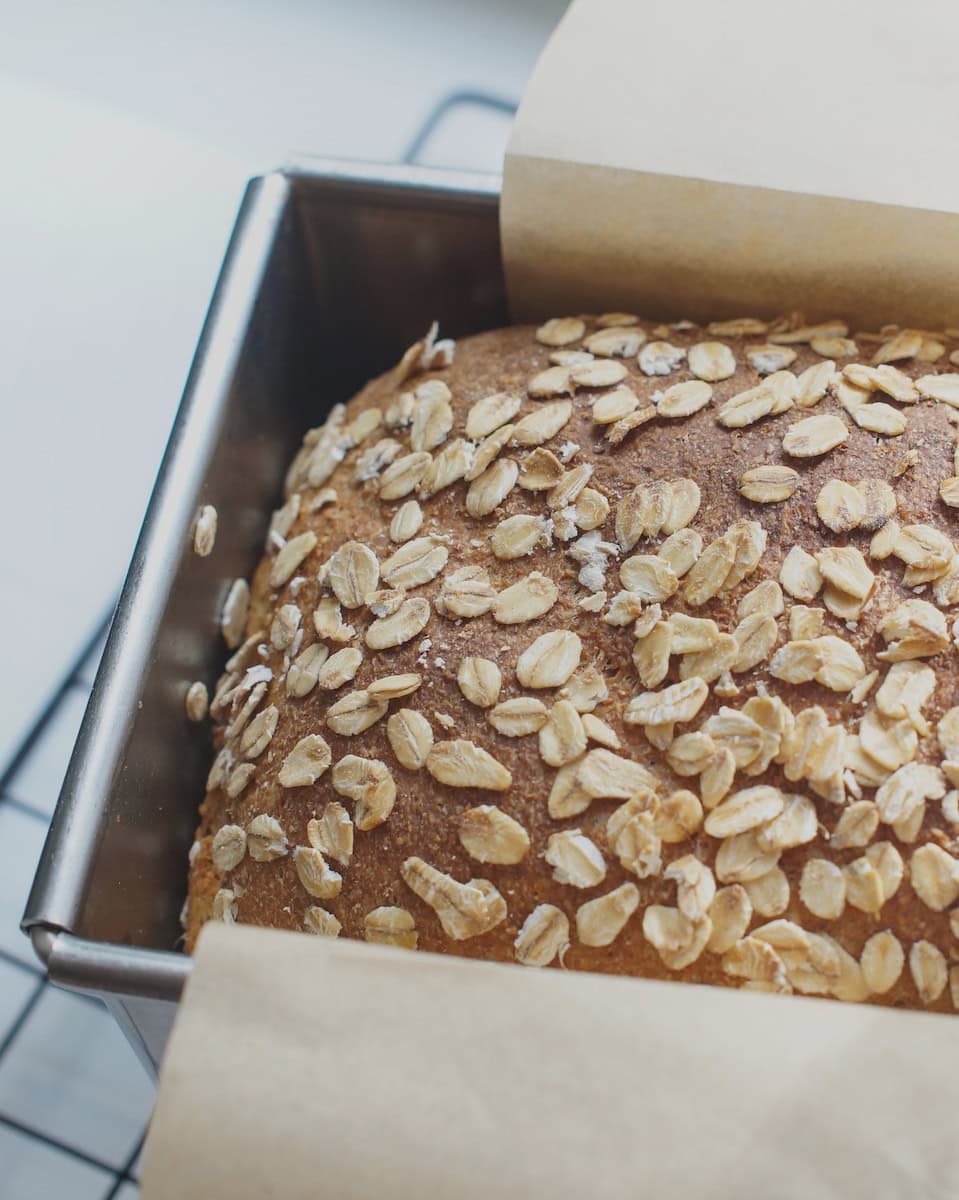 Closeup of finished toasted oat and honey discard sandwich bread, baked to golden perfection. The bread rests in a silver pullman loaf pan on a black cooling rack in bright sunlight.