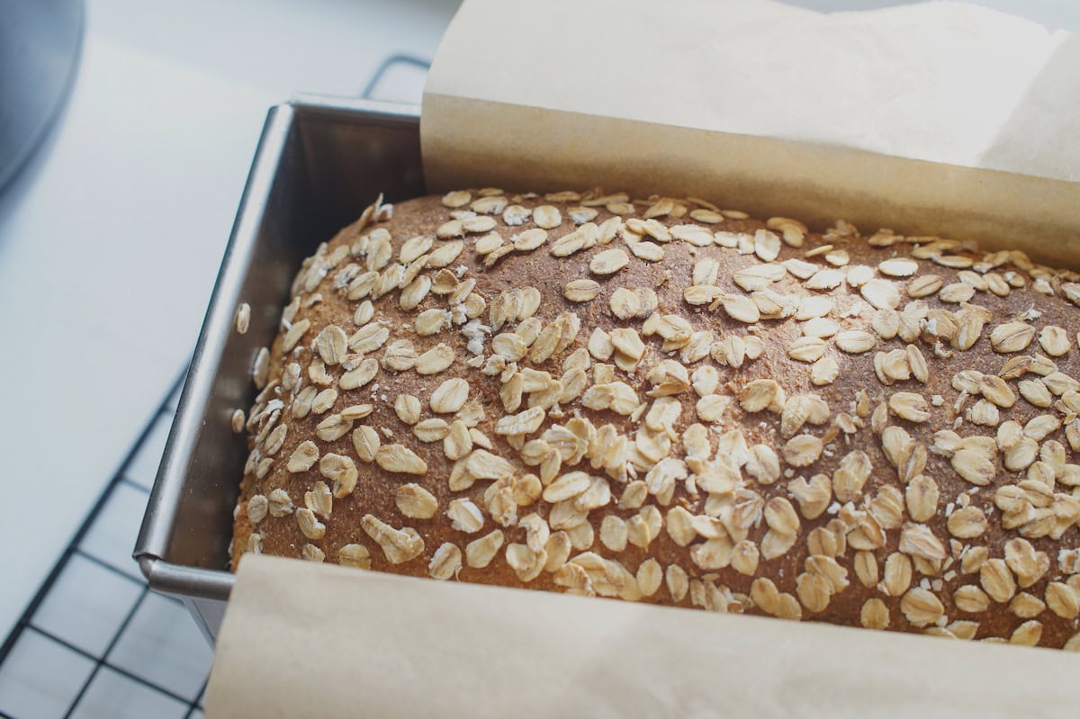 Closeup of finished toasted oat and honey discard sandwich bread, baked to golden perfection. The bread rests in a silver pullman loaf pan on a black cooling rack in bright sunlight.