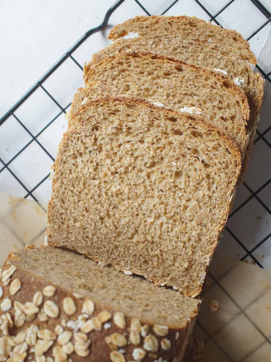 Slices of tender oat and honey whole wheat discard sandwich bread resting next to the loaf on a black wire rack. You can see the toasty oat crust and the fluffy crumb.
