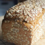 Closeup of finished toasted oat and honey discard sandwich bread, baked to golden perfection. The whole wheat bread rests on a black cooling rack in bright sunlight.