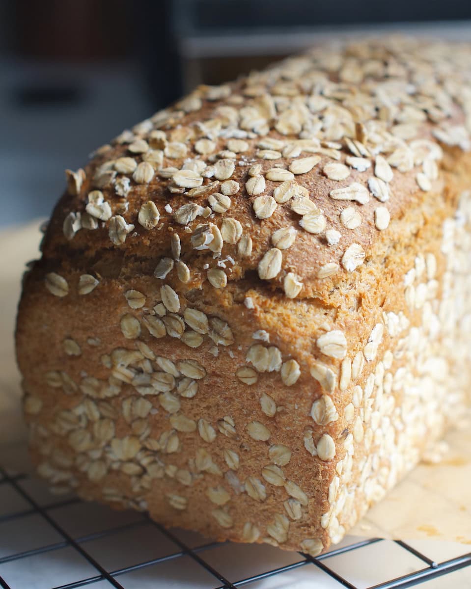 Closeup of finished toasted oat and honey discard sandwich bread, baked to golden perfection. The whole wheat bread rests on a black cooling rack in bright sunlight.