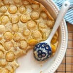 Golden yellow mirabelle einkorn clafoutis cooling off on a black wire rack. The pan is surrounded by a green striped tea towel and a patterned blue ceramic serving spoon rests in the pan, where a serving has already been spooned out.