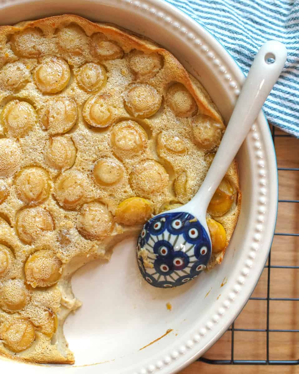 Golden yellow mirabelle einkorn clafoutis cooling off on a black wire rack. The pan is surrounded by a green striped tea towel and a patterned blue ceramic serving spoon rests in the pan, where a serving has already been spooned out.