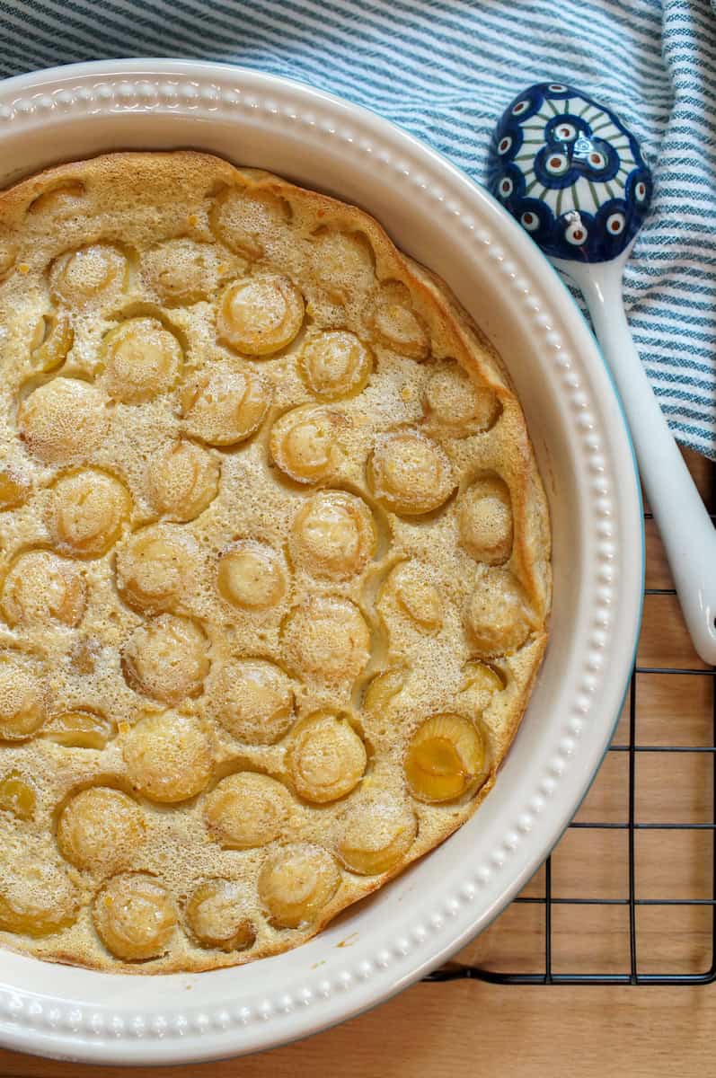 Golden yellow mirabelle einkorn clafoutis cooling off on a black wire rack. The pan is surrounded by a green striped tea towel and a patterned blue ceramic serving spoon.