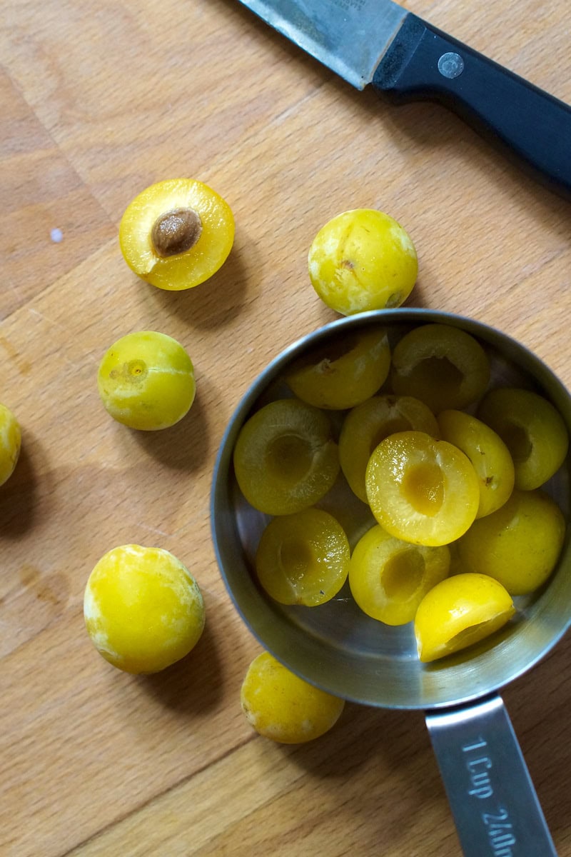 Vibrant yellow green mirabelle plums, sliced in a 1-cup measure. Some are halved, some whole, one shows the tiny stone in the middle.