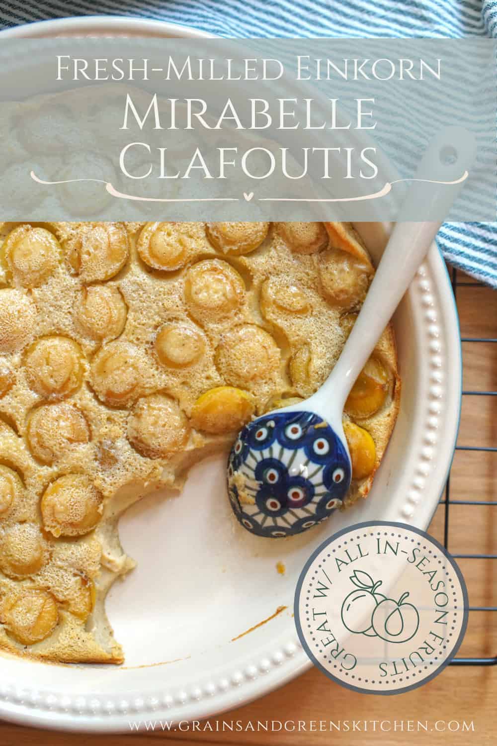 Golden yellow mirabelle einkorn clafoutis cooling off on a black wire rack. The pan is surrounded by a green striped tea towel and a patterned blue ceramic serving spoon rests in the pan, where a serving has already been spooned out. On-image title: "Mirabelle Einkorn Clafoutis, Fresh Flour Optional"