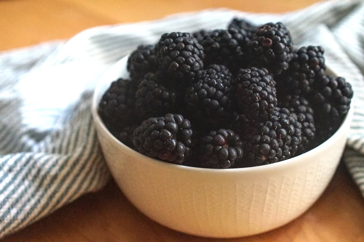 Closeup of glossy blackberries in a patterned white bowl, draped with a striped tea towel.